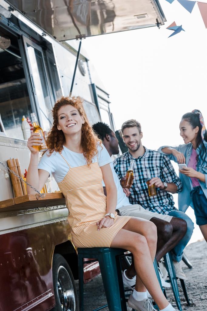 feliz pelirroja chica celebración botella con cerveza cerca multicultural amigos y comida camión
  - Foto, Imagen