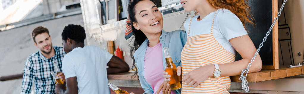 tiro panorâmico de meninas felizes segurando garrafas com cerveja perto de homens multiculturais e caminhão de comida
  - Foto, Imagem