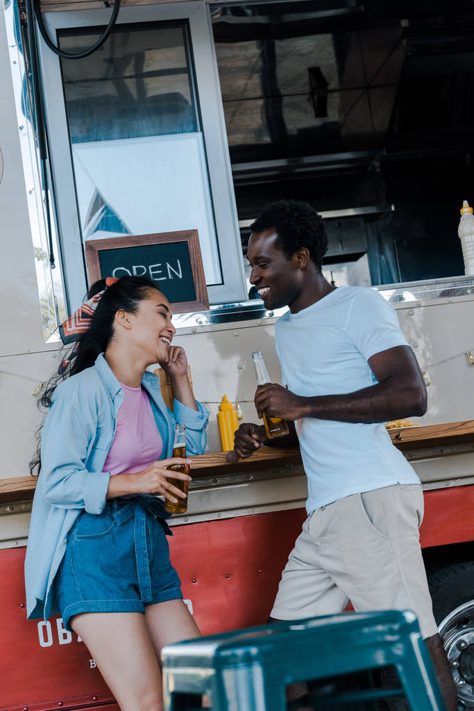ビールのボトルを持つアフリカ系アメリカ人男性の近くで微笑む陽気なアジアの女の子  - 写真・画像