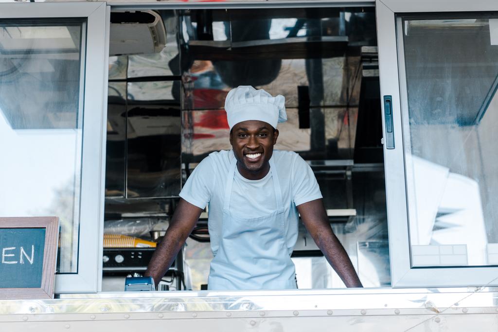 シェフのユニフォームと帽子のフードトラックから笑顔の陽気なアフリカ系アメリカ人の男性  - 写真・画像