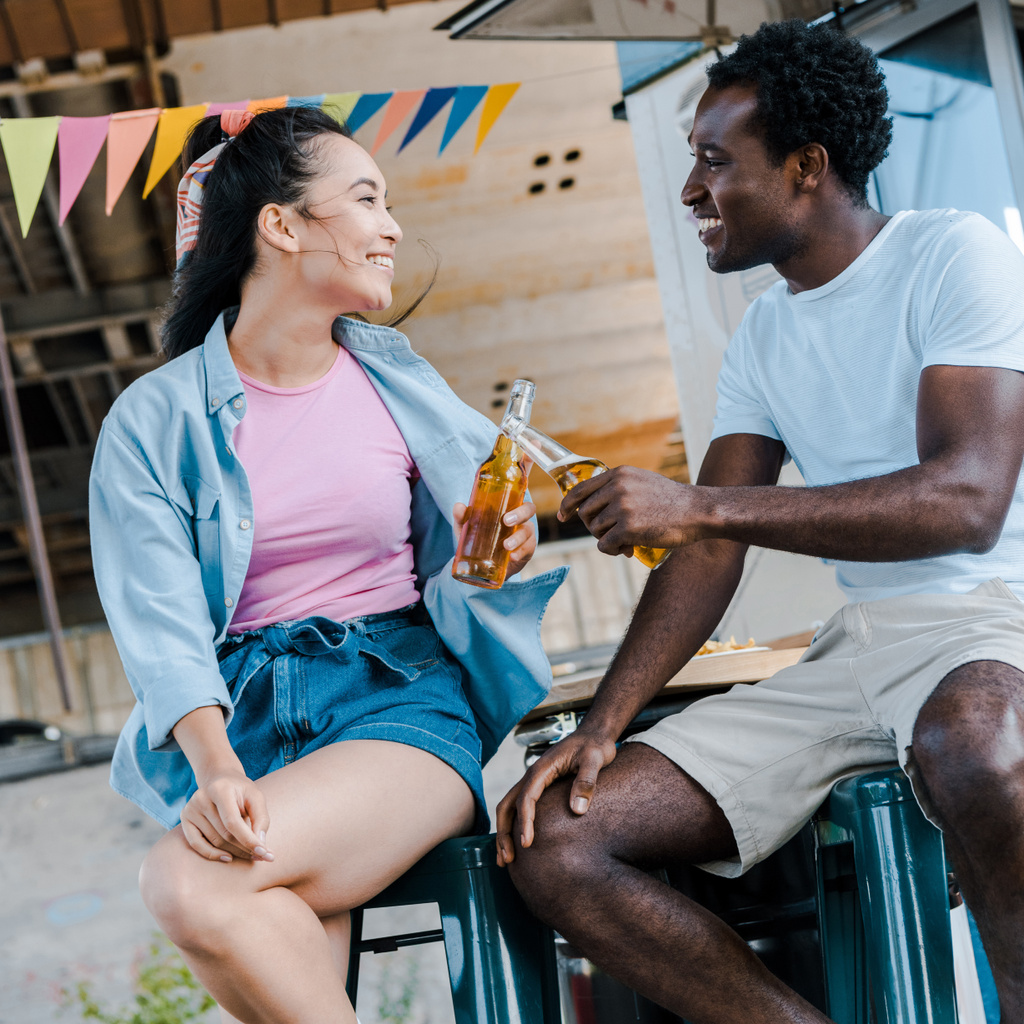 陽気なアジアの女性は、アフリカ系アメリカ人の男性の近くに微笑み、フードトラックの近くでビールのボトルをクリンキング  - 写真・画像