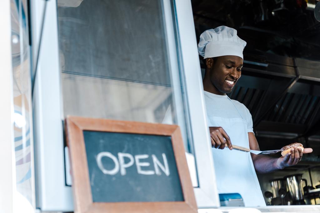 foco seletivo de feliz afrian americano homem segurando espátula enquanto prepara hambúrguer perto de giz bordo com letras abertas
  - Foto, Imagem