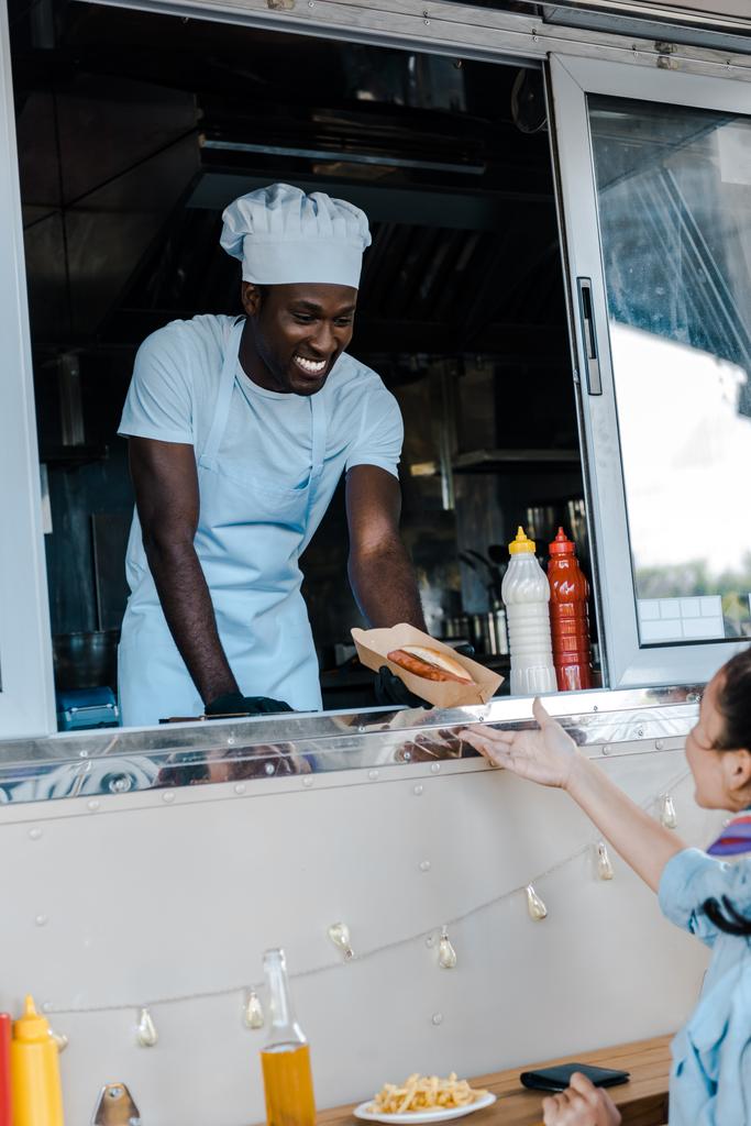 selektiver Fokus des fröhlichen afrikanisch-amerikanischen Mannes, der dem Kunden einen Kartonteller mit Pommes frites schenkt  - Foto, Bild