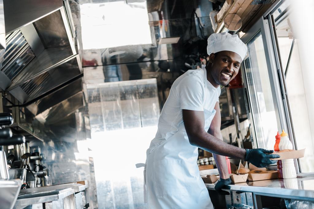 selectieve focus van vrolijke Afro-Amerikaanse chef-kok glimlachend karton plaat in Food truck  - Foto, afbeelding