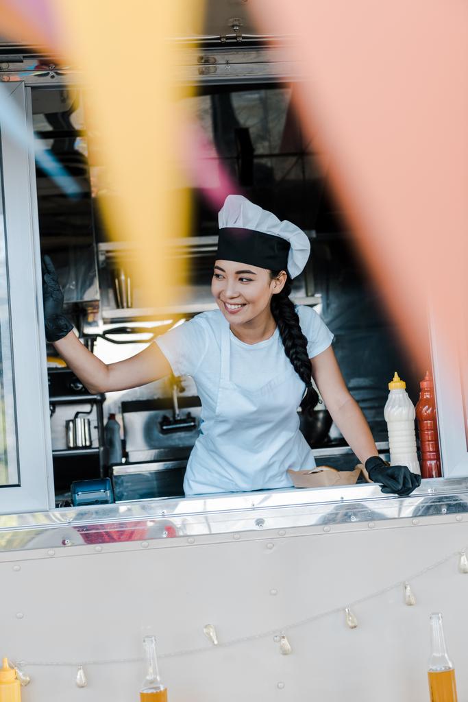 フードトラックで笑顔の帽子とシェフ制服で陽気なアジアの女性の選択的な焦点  - 写真・画像
