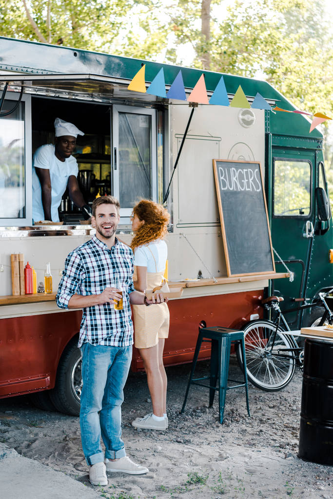 счастливый мужчина держит бутылку с пивом рядом с женщиной и африканским американцем в фургоне с едой
  - Фото, изображение