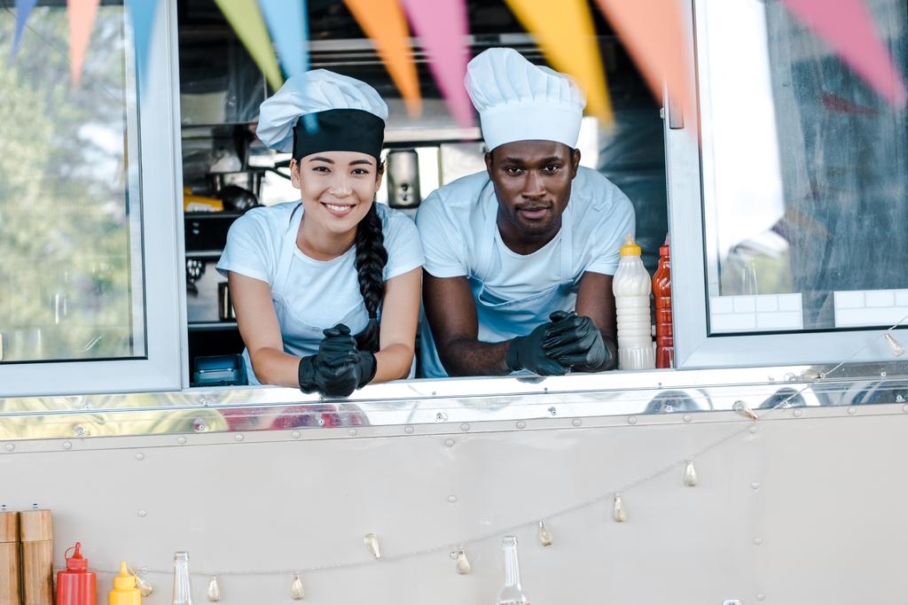 フードトラックから微笑む帽子をかぶったアジアの女の子とアフリカ系アメリカ人男性の選択的な焦点  - 写真・画像