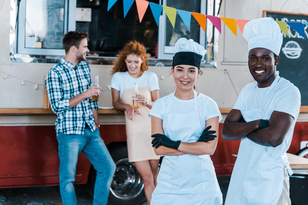 εκλεκτική εστίαση των χαρούμενα πολυπολιτισμικού σεφ χαμογελώντας κοντά σε φορτηγό τροφίμων και τους πελάτες  - Φωτογραφία, εικόνα