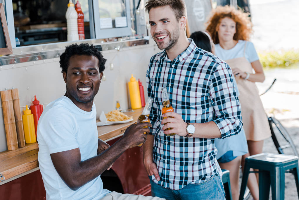вибірковий фокус веселих мультикультурних чоловіків, що тримають пляшки пива біля дівчат та харчової вантажівки
  - Фото, зображення