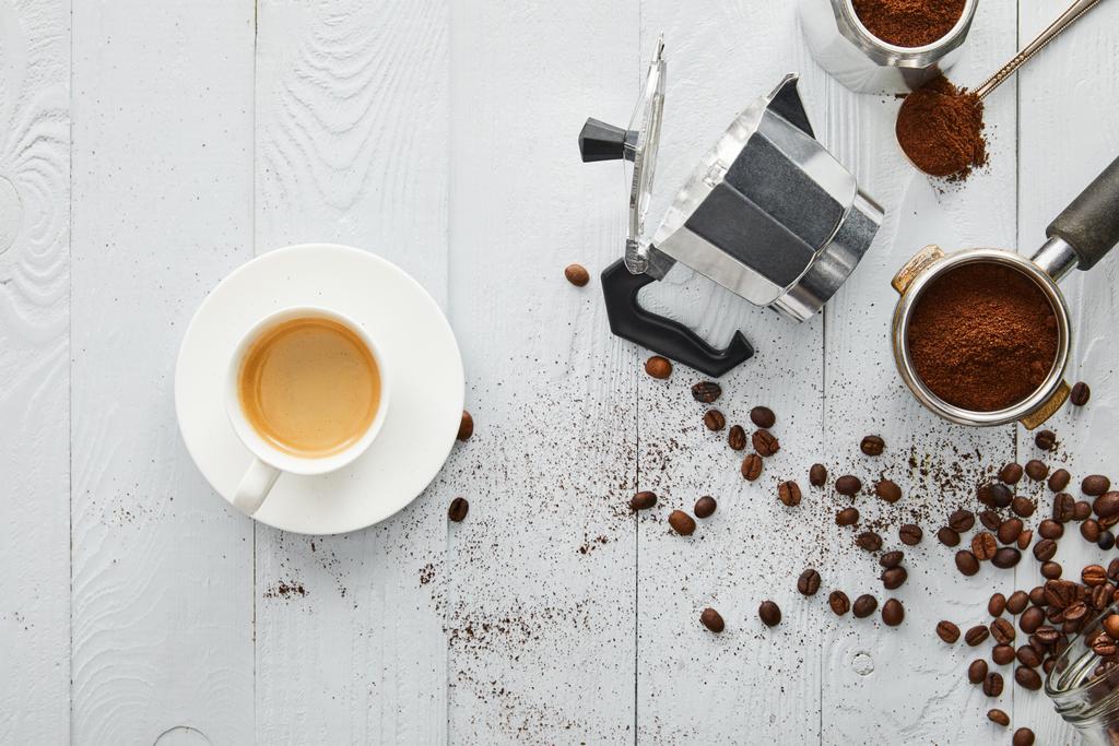 Вид зверху чашки кави на блюдці біля кавоварки гейзера, портативний фільтр і ложка на білій дерев'яній поверхні з кавовими зернами
 - Фото, зображення