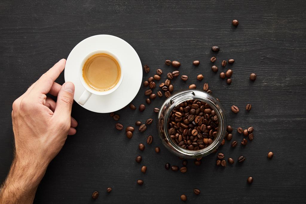 Вид зверху чоловічої руки з чашкою кави біля скляної банки з кавовими зернами
 - Фото, зображення