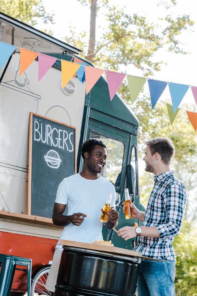cheerful multicultural friends holding bottles of beer near french fries and food truck  - Photo, Image