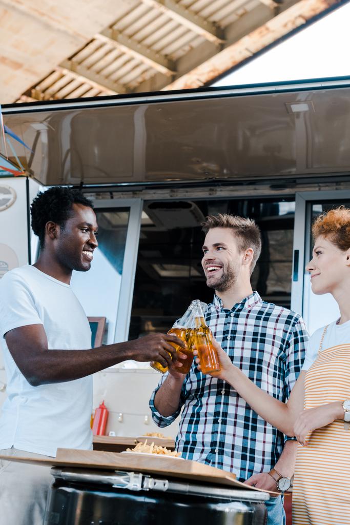 επιλεκτική εστίαση των πολυπολιτισμικού άνδρες που Κουδουνά μπουκάλια μπύρας με ευτυχισμένη κοκκινομάλλα γυναίκα κοντά στο φορτηγό τροφίμων - Φωτογραφία, εικόνα