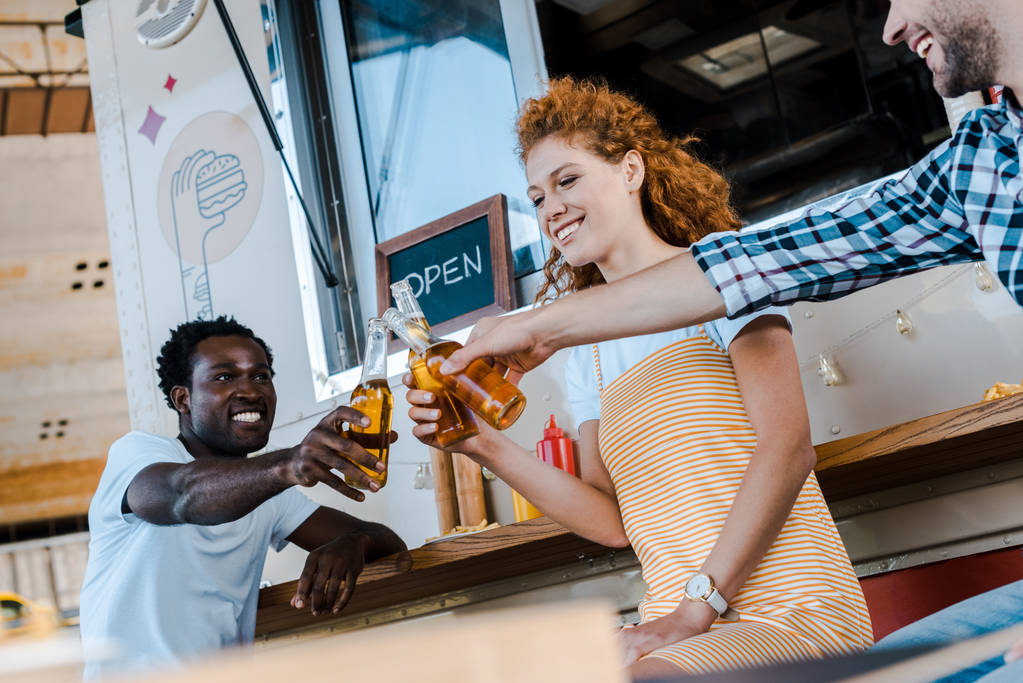 komea monikulttuurinen miehet clinking pulloa olutta houkutteleva punapää nainen lähellä ruoka kuorma
  - Valokuva, kuva