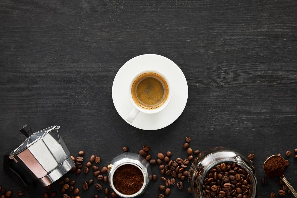 Bovenaanzicht van kopje espresso op schotel in de buurt van geiser koffie maker, glazen pot en lepel op donker houten oppervlak met koffiebonen - Foto, afbeelding