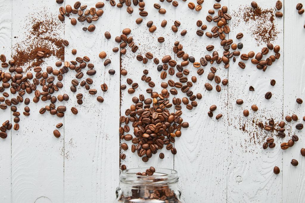 ガラス瓶で白い木製の表面に散らばったコーヒー豆 - 写真・画像