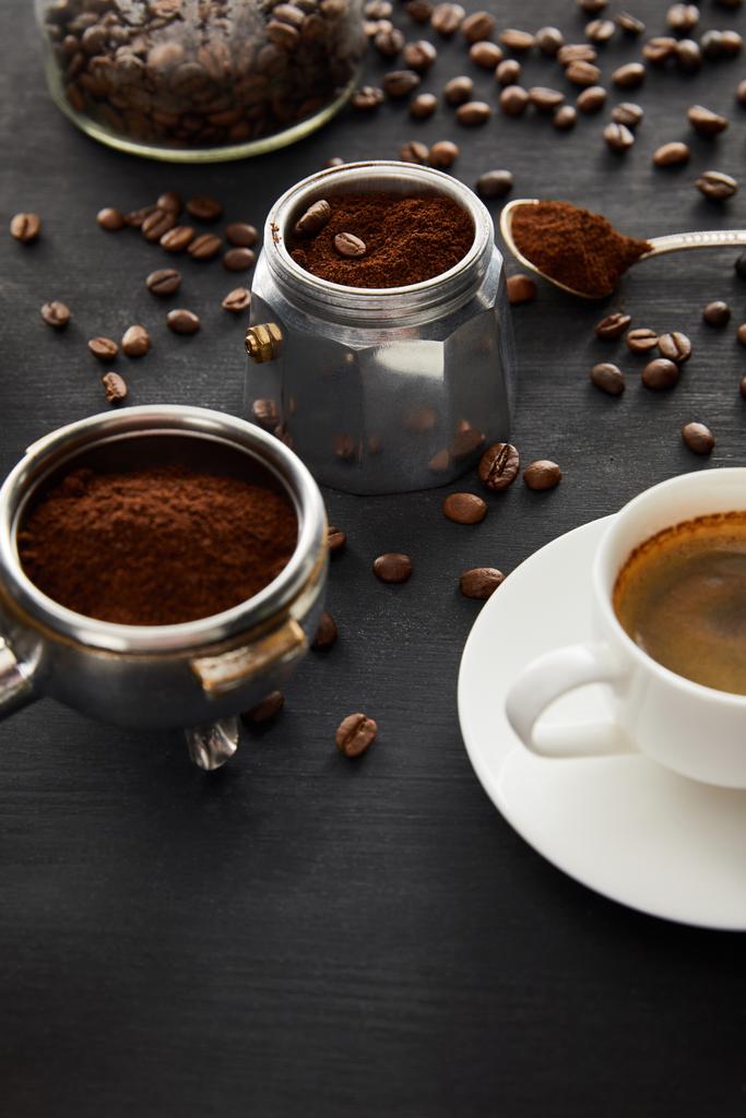 Onderdeel van geiser koffie maker in de buurt van kopje koffie op schotel, filterhouder en lepel op donker houten oppervlak met koffiebonen - Foto, afbeelding
