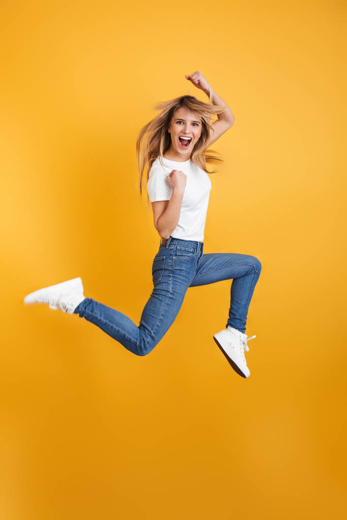 Эмоциональный крик положительный молодой блондинки прыжки изолированные через желтый фон стены, одетые в белые повседневные футболки сделать жест победителя
. - Фото, изображение