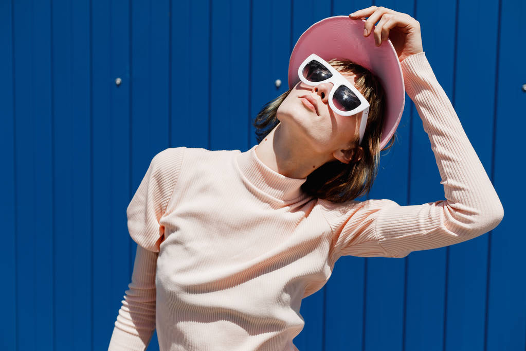 Chica con estilo en un vestido suavemente rosa, gafas de sol blancas y visera rosa posa contra una pared de metal azul brillante en el exterior en un día soleado
 - Foto, imagen