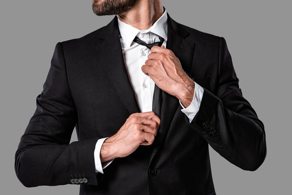 oříznutý pohled na stylový pohledný obchodník v černém obleku dotyk kravatu izolovaný na šedé - Fotografie, Obrázek