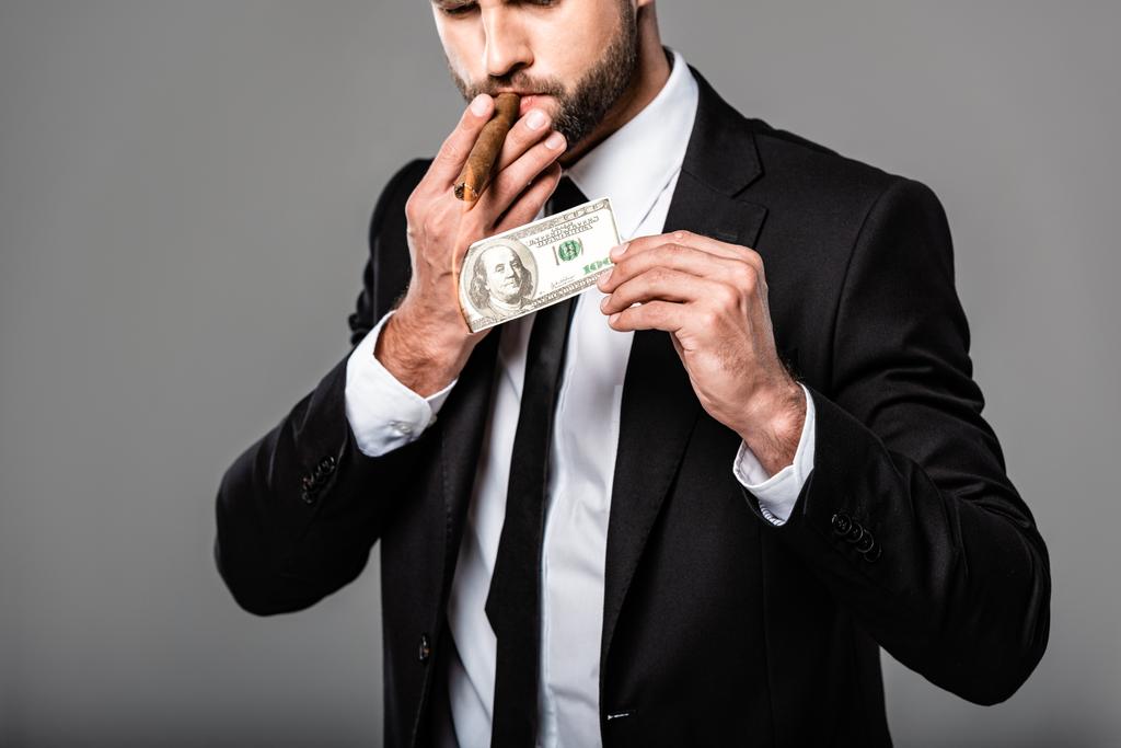 abgeschnittene Ansicht des reichen erfolgreichen Geschäftsmannes im schwarzen Anzug, der Zigarre aus brennender Dollarnote isoliert auf grau anzündet - Foto, Bild