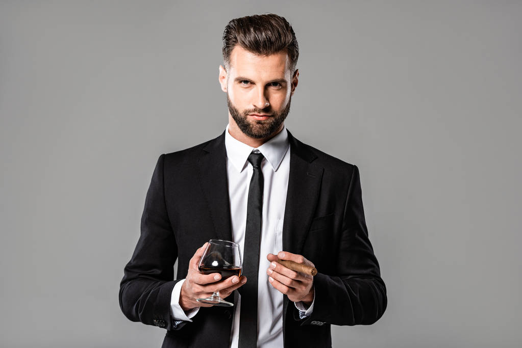 επιτυχημένος επιχειρηματίας με μαύρο κοστούμι κρατώντας γυαλί με ουίσκι και πούρο απομονώθηκαν σε γκρι - Φωτογραφία, εικόνα