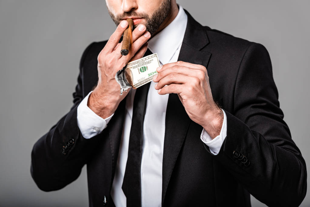 частичный взгляд богатого успешного бизнесмена в черном костюме, освещающего сигару от горящей долларовой купюры, изолированной на сером
 - Фото, изображение