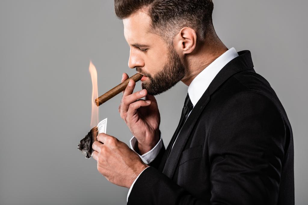 Zijaanzicht van de rijke succesvolle zakenman verlichting van sigaar van brandende dollar bankbiljet geïsoleerd op grijs - Foto, afbeelding