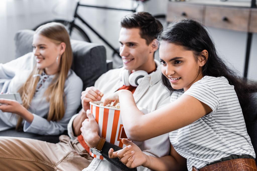 multicultural friends sitting on sofa and eating popcorn in apartment  - Photo, Image