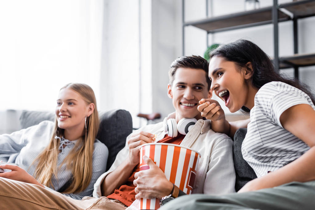 wielokulturowych przyjaciół siedzi na kanapie i jedzenia popcorn w mieszkaniu  - Zdjęcie, obraz