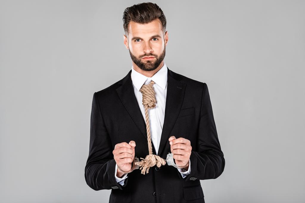 handsome businessman in black suit and handcuffs with noose on neck isolated on grey - Photo, Image