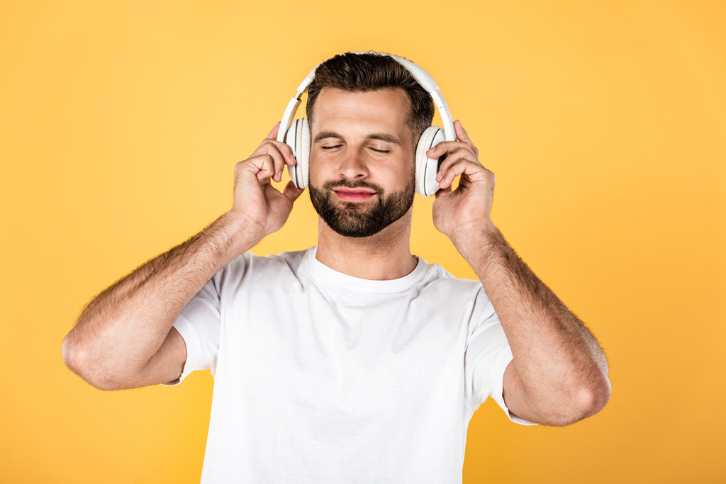 glücklicher gutaussehender Mann in weißem T-Shirt, der Musik mit Kopfhörern hört, mit geschlossenen Augen, isoliert auf gelb - Foto, Bild
