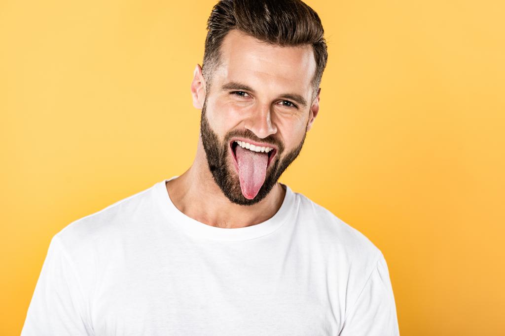 handsome funny man in white t-shirt showing tongue isolated on yellow - Photo, Image