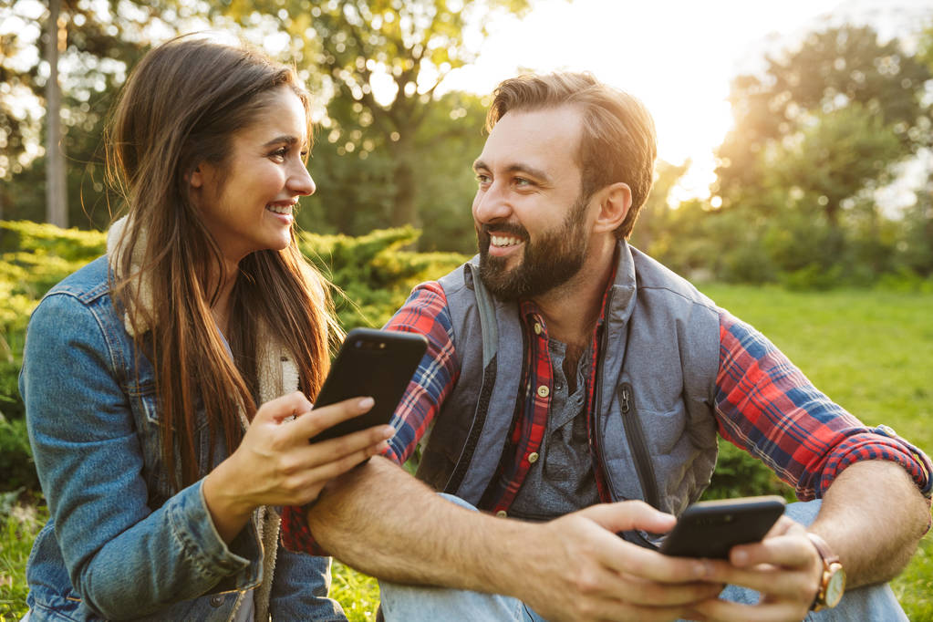 Image de couple joyeux homme et femme utilisant des téléphones cellulaires et se regardant tout en se reposant dans un parc verdoyant
 - Photo, image