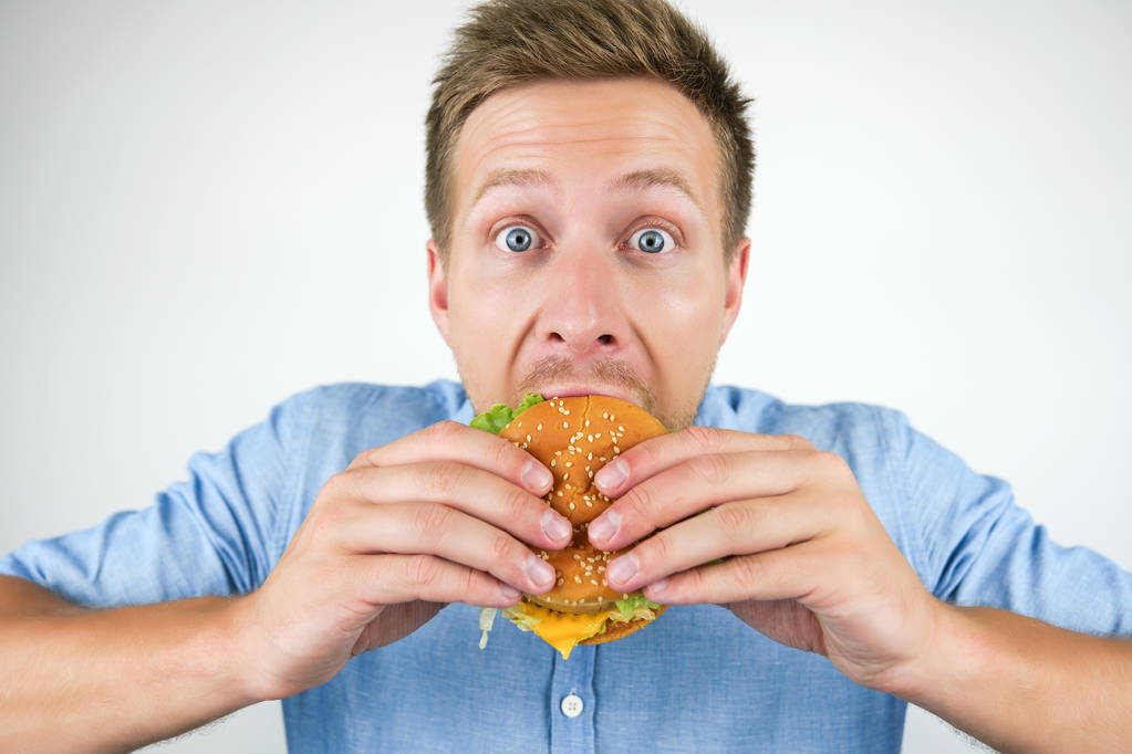 junger schöner Mann beißt Cheeseburger aus Fast-Food-Restaurant hungrig auf isoliertem weißen Hintergrund - Foto, Bild