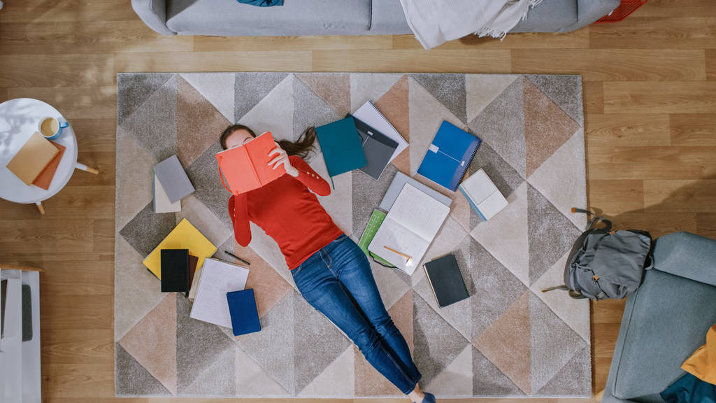 Jong meisje in rode vacht en blauwe jeans ligt op een vloer, het lezen van een notebook. Kijkt boven, glimlacht en lacht. Gezellige woonkamer met modern interieur met tapijt, werkmappen en rugzak. Top View. - Foto, afbeelding