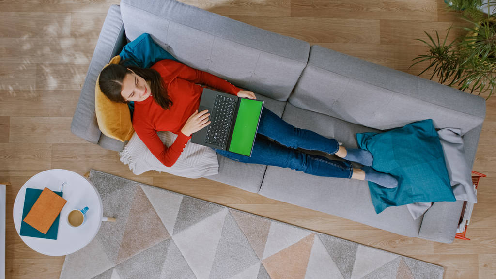 junges Mädchen in rotem Mantel und blauer Jeans legt sich auf ein Sofa und arbeitet an einem Laptop mit grünem Bildschirm. gemütliches Wohnzimmer mit modernem Interieur mit Teppich, Pflanzen, Couchtisch und Holzboden. von oben nach unten - Foto, Bild