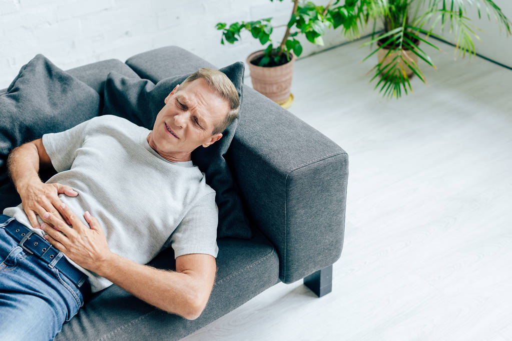 όμορφος άντρας με μπλουζάκι με στομαχόπονο που βρίσκεται στον καναπέ  - Φωτογραφία, εικόνα
