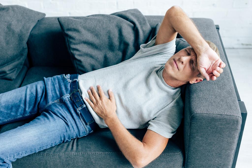 handsome man in t-shirt with stomachache lying on sofa  - Photo, Image