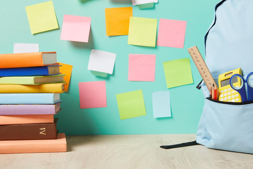 blue backpack with supplies in pocket near books and multicolored sticky notes on turquoise wall - Photo, Image