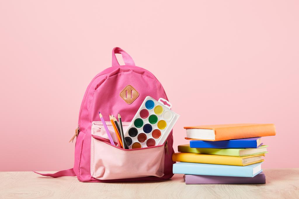 mochila rosa con suministros en el bolsillo cerca de libros de colores aislados en rosa
 - Foto, Imagen