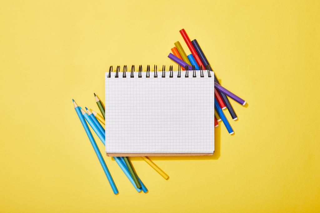 Widok z góry kolorowych ołówków i długopisów rozrzuconych w pobliżu pustego notebooka na żółtym - Zdjęcie, obraz