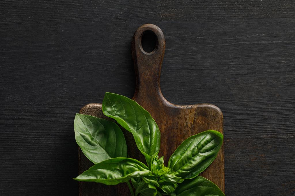Top view of brown wooden cutting board with basil twig on dark surface - Photo, Image
