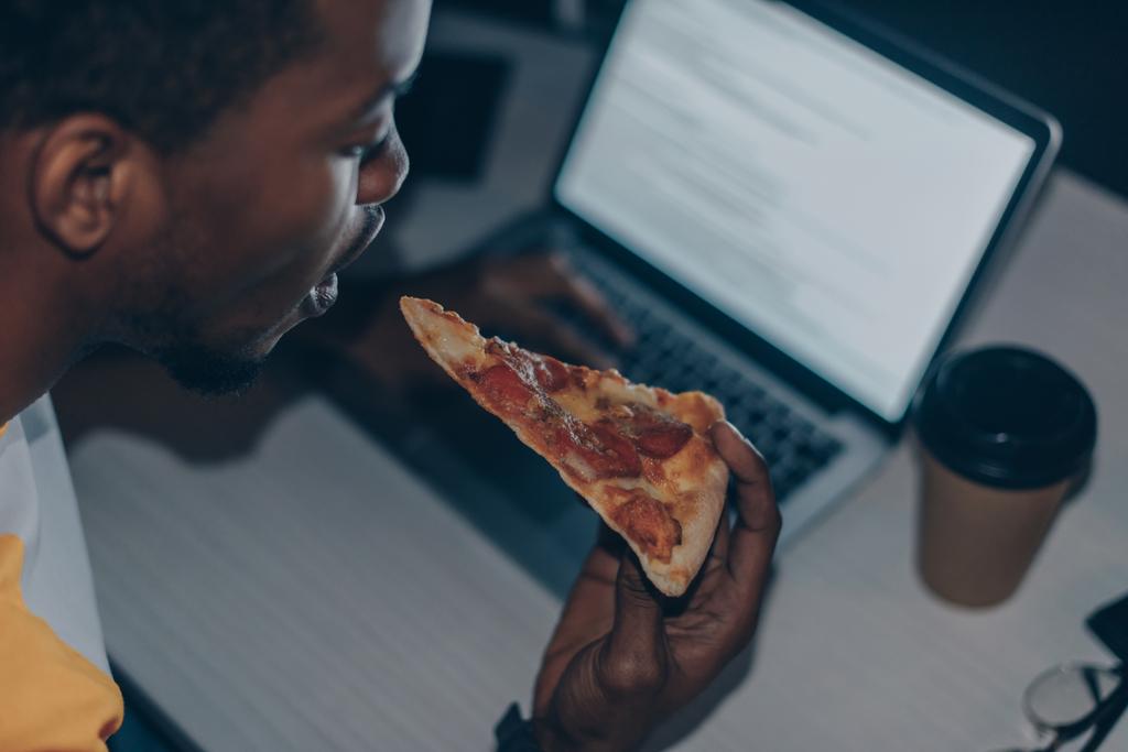 若いアフリカ系アメリカ人プログラマーがオフィスで夜働きながらピザを食べる - 写真・画像