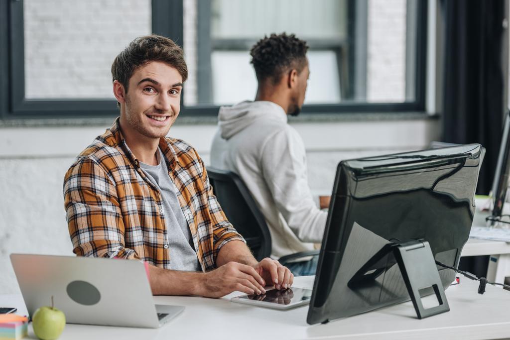 cheerful programmer smiling at camera while sitting near african american programmer - Photo, Image