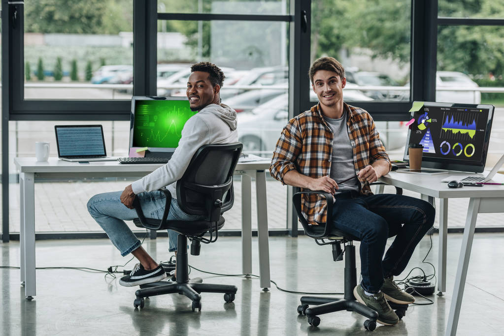 zwei junge multikulturelle Programmierer blicken in die Kamera, während sie neben Computermonitoren sitzen, auf denen Grafiken und Diagramme auf dem Bildschirm zu sehen sind - Foto, Bild