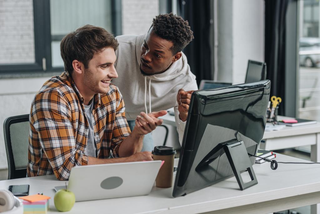 dos programadores multiculturales sonrientes apuntando al monitor de la computadora mientras trabajan juntos en la oficina
 - Foto, Imagen