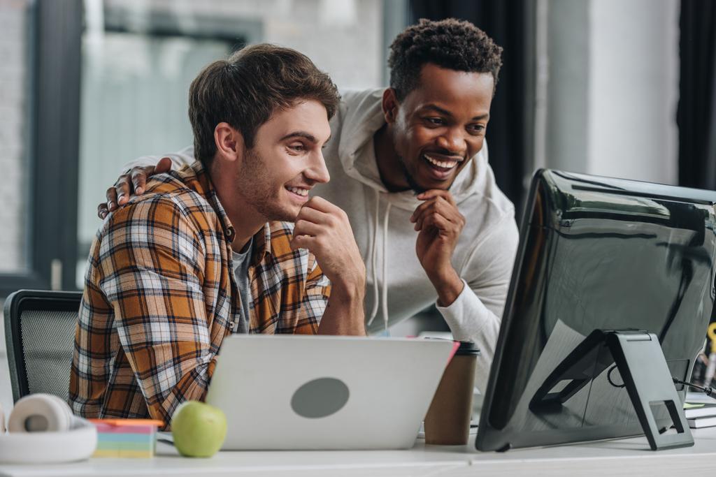 dois programadores multiculturais alegres que olham para o monitor do computador enquanto trabalham no escritório
 - Foto, Imagem