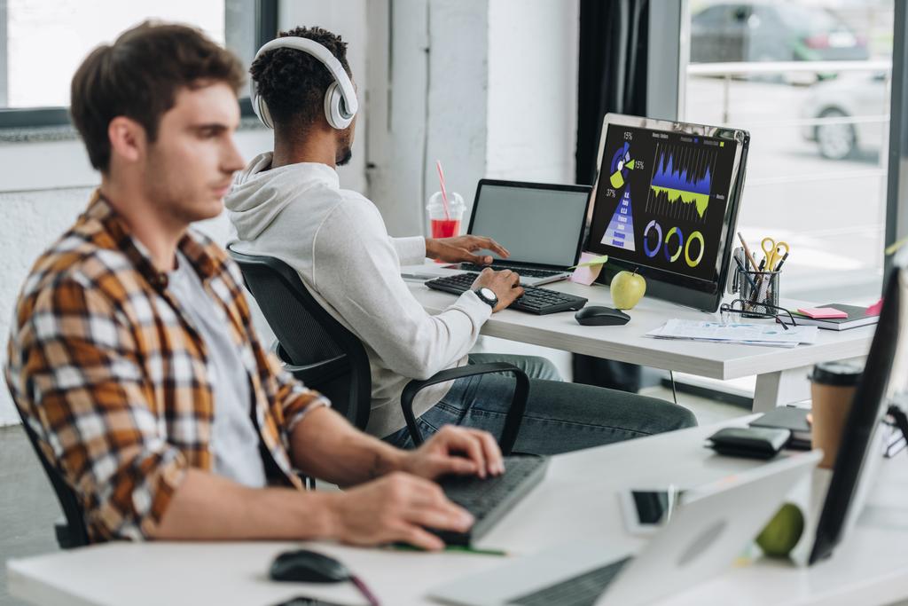 ofiste meslektaşının yanında ekranda grafikler ve grafikler ile bilgisayar monitörü yakınında oturan kulaklık afro-amerikan programcı nın arka görünümü - Fotoğraf, Görsel