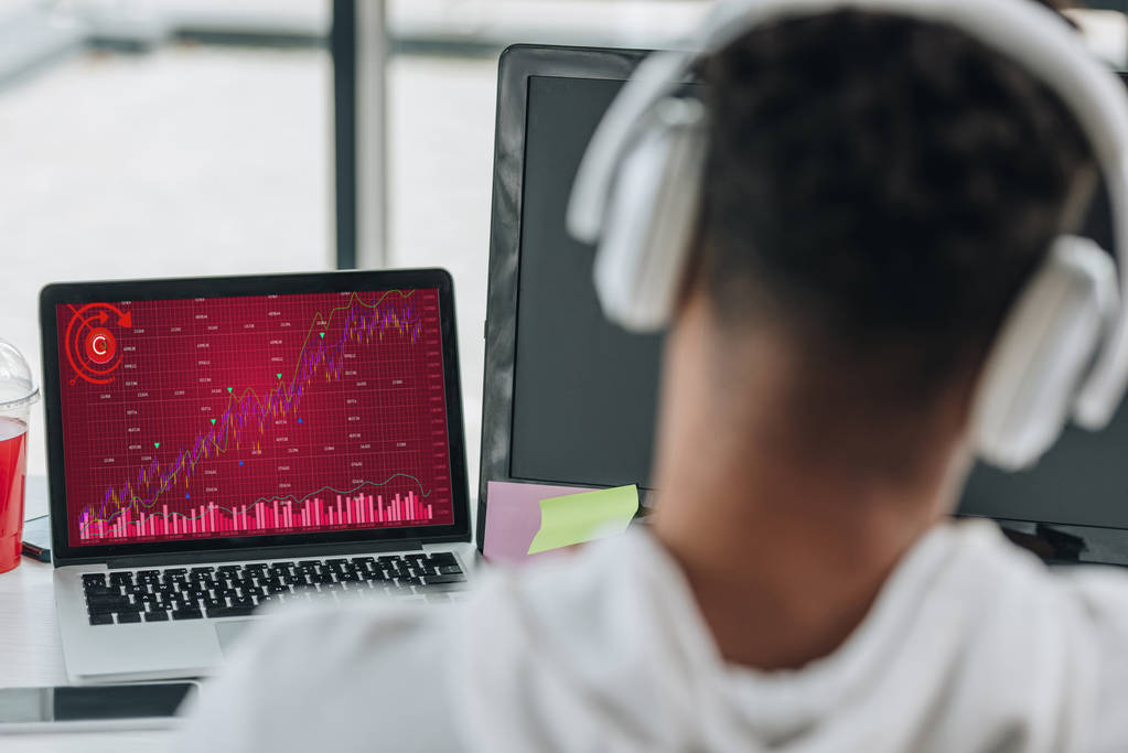 Widok z tyłu afrykańskiego amerykańskiego programisty w słuchawkach patrząc na laptopa z wykresów i wykresów na ekranie - Zdjęcie, obraz